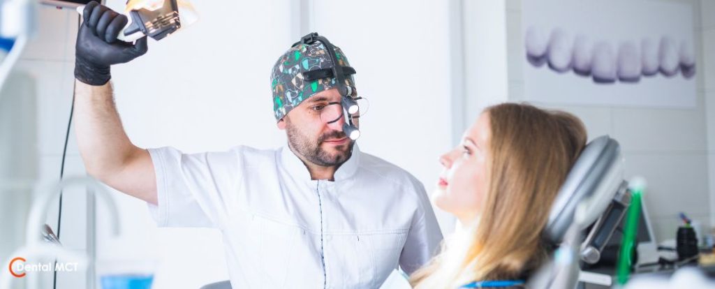 מה ההבדל בין מומחה פה ולסת לרופא שיניים כללי? - רופא שיניים עם כובע צבעוני מאיר על מטופלת בלונדינית ששוכבת על כיסא טיפולים במרפאה. בצד שמאל למטה לוגו אתר dentalmct